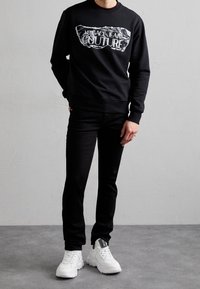 Svart sweatshirt med grafiskt tryck, kombinerad med svarta smala jeans och vita sneakers. Sweatshirten har rund halsringning och ribbade muddar.