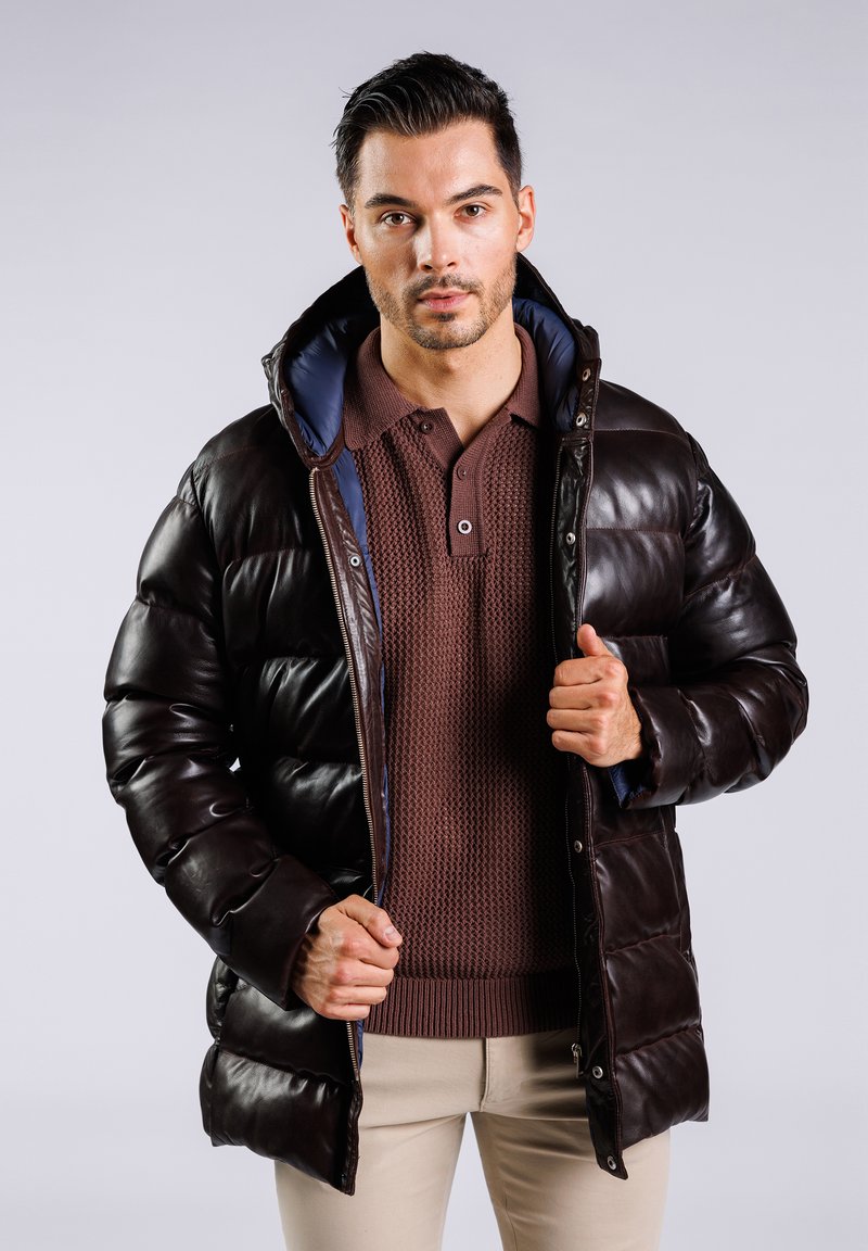 Chaqueta de plumón marrón con un acabado brillante, que cuenta con capucha y forro azul. Lleva puesta abierta sobre una camiseta de polo marrón texturizada.