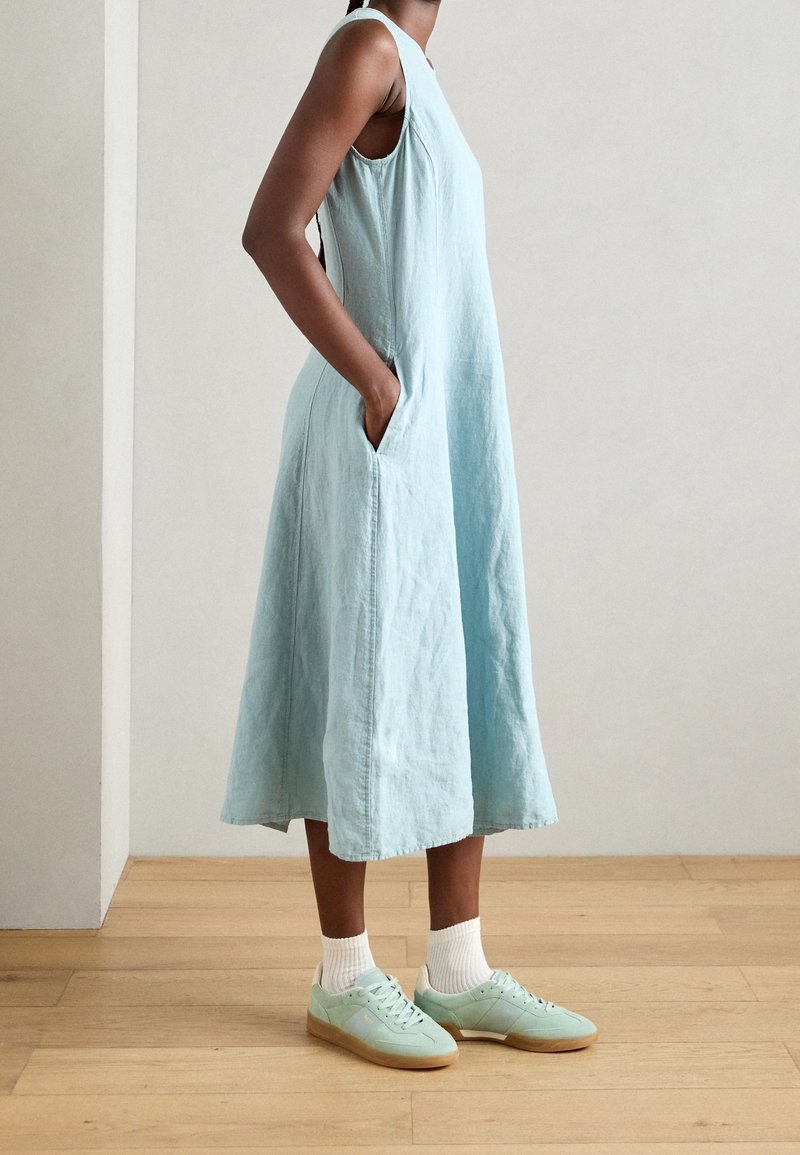 Femme portant une robe midi bleu clair sans manches avec poches, des chaussettes blanches et des baskets vert clair, debout sur un sol en bois.