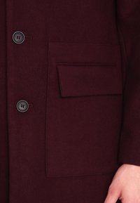 Bordeauxroter Wollmantel mit Knopfverschluss, oberer Tasche und weicher Textur. Der Mantel hat eine glatte Oberfläche und ein klassisches Design.