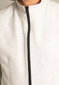 Ljusgrå fleecejacka med hög krage och svart dragkedja. Tyget har en texturerad yta och en slät finish längs dragkedjan.