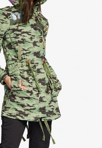 Kamouflage-mönstrad militärjacka i gröna nyanser, med broderade fjärilar, dragsko i midjan och knäppning med knappar.