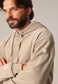 Beige geribbelde hoodie met een trekkoordkap, voorzien van een klein logo-patch op de mouw en accentdetails in een lichtere kleur.