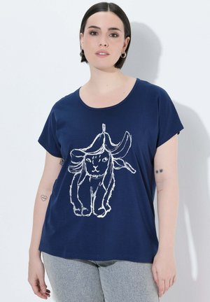 Kobieta z krótkimi ciemnymi włosami, ubrana w granatowy t-shirt z białą liniową grafiką kota pokrytego skórkami od banana, w połączeniu z jasnoszarymi spodniami.