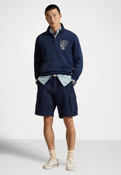Tumši zils džemperis ar puszipa apkakli un logotipu, pārģērbts pāri gaiši zilai džinsa krekliņai, papildināts ar tumši ziliem cargo šortiem un baltām sporta kurpēm.