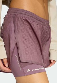 Lagane ljubičaste hlače s višeslojnim dizajnom, perforiranim detaljima za ventilaciju i elastičnim pojasom. Prikazan je logo marke.