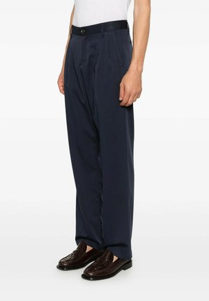 Uomo che indossa pantaloni plissettati blu scuro e mocassini in pelle marrone scuro, in piedi con una mano rilassata al fianco.