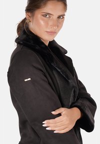 Cappotto in pelliccia sintetica nera con colletto ampio, caratterizzato da un logo dorato sulla manica. Il tessuto ha una texture liscia e un design elegante.