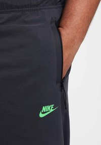Pantaloni sportivi neri Nike con tasca con zip, caratterizzati da un logo verde neon. Il tessuto è liscio con una finitura opaca e una vita elastica.