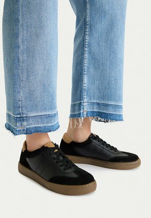 Czarne trampki z brązowymi podeszwami noszone z podwiniętymi, jasnoniebieskimi, postrzępionymi dżinsami na osobie stojącej na białym tle.