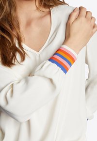 Hvid top med V-hals og voluminøse ærmer, prydet med en flerfarvet stribet manchet i pink, orange, blå og lilla.