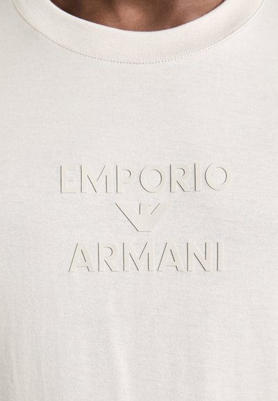 Ανοιχτό μπεζ βαμβακερό t-shirt με υφασμένο, ανάγλυφο λογότυπο "EMPORIO ARMANI" στο στήθος. Στρογγυλή λαιμόκοψη και κοντά μανίκια.
