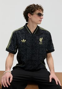 Fekete rövid ujjú ing mintával, sárga Adidas és Liverpool FC logókkal. Bordázott gallér és szegély; texturált anyag, laza szabás.