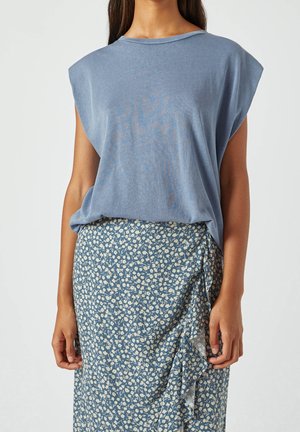 Kvinde iført en løs ærmeløs blå top, stoppet ned i et højtaljet blåt skørt med hvidt blomstermønster og detaljer med flæser.