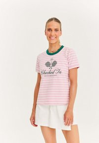 Kobieta ubrana w różową i białą koszulkę w paski z napisem "Zameldowana, wyłączona" oraz białą spódnicę, uśmiechająca się na tle jednolitym.