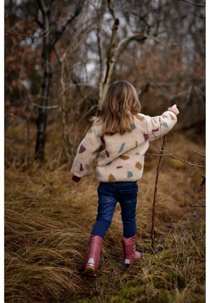 Enfant portant une veste polaire crème avec des formes abstraites multicolores, un jean bleu et des bottes en caoutchouc roses, marchant à travers une haute herbe et des arbres.
