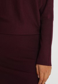 Primo piano di un braccio e una mano di una persona che indossa un vestito a maniche lunghe fitted in maglia a coste di un intenso bordeaux.