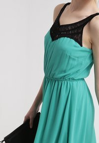 Tyrkysové šaty s černým krajkovým korzetem se geometrickým vzorem, s nastavitelnými ramínky a elastickým pasem, doplněné černou kabelkou.