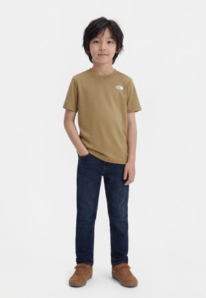 Boy with dark hair stands facing forward wearing a brown t-shirt, dark blue jeans, and brown sneakers against a white background.