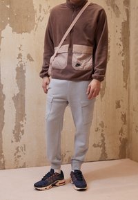 Haut en polaire marron à fermeture éclair quart avec deux poches à rabat roses, associé à un pantalon de jogging gris clair avec des poches latérales. Logo Nike noir sur les deux pièces.