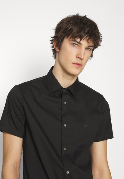 Chemise noire à manches courtes avec boutons, fabriquée en tissu lisse, dotée d'un col pointu, présentant un logo tonal sur la poitrine et des boutons argentés.