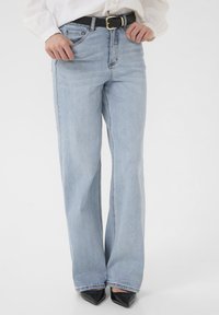 Person, die helle blaue, weite Jeans, einen schwarzen Gürtel, ein weißes Hemd und schwarze spitze High Heels trägt, steht vor einem weißen Hintergrund.