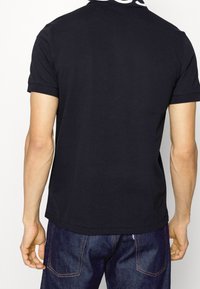 Czarna koszulka polo z teksturowanym kołnierzykiem, krótkimi rękawami i prostym dołem. Noszona z ciemnymi dżinsami, które mają minimalne detale wzornicze.