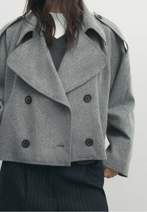 Femme portant un manteau court en laine grise à double boutonnage sur un pantalon sombre à fines rayures et un haut blanc et gris superposé.