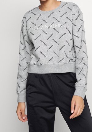 Persona con una sudadera gris de Calvin Klein con logotipos negros repetidos y un gran logo blanco en el pecho, combinada con pantalones negros.