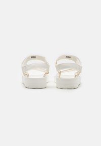 Valkoiset sandaalit, joissa on litteat pohjat, kestävästä kankaasta valmistetut hihnat ja säädettävät tarranauhat, minimalistisella muotoilulla.