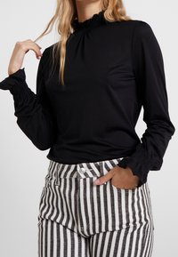 Schwarzes Langarmoberteil mit gerafftem Ausschnitt und gerafften Bündchen, kombiniert mit hoch taillierten, schwarz-weißen gestreiften Hosen mit glitzerndem Finish.