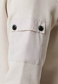 Felpa color beige chiaro con un tessuto testurizzato, caratterizzata da una tasca liscia a contrasto sulla manica fissata da bottoni a pressione neri.
