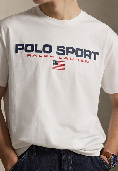 Λευκό βαμβακερό μπλουζάκι με τις λέξεις «POLO SPORT» και «RALPH LAUREN» τυπωμένες σε μπλε και κόκκινο. Διαθέτει μικρό γραφικό της αμερικανικής σημαίας. Άνετη εφαρμογή.