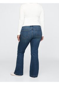 Jeans i mörkblå tvätt med utsvängd benen. Har diskreta sömmar på bakfickorna och en slät textur. Parat med en vit långärmad topp.