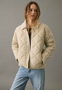 Mujer joven con cabello ondulado que lleva una chaqueta de microfibra beige con cuello de sherpa y jeans azul claro, de pie con las manos en los bolsillos.