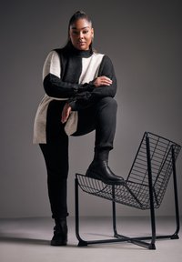 Czarny i biały sweter w paski o fakturowanym splotcie, czarne spodnie i czarne botki, pozujący z jedną stopą na metalowej koszyczkowej konstrukcji.