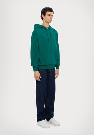 Felpa verde in cotone con coulisse regolabile, tasca frontale e polsini a costine, abbinata a jeans dritti blu scuro e sneakers bianche.