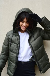 Mörk olivfärgad pufferjacka med huva, med ett quiltat mönster, framzipper och dragsko. Bärs över en vit skjorta och svarta jeans.