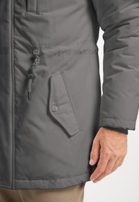 Manteau isolé gris avec une poche latérale à fermeture pression et un cordon de serrage en accent, présentant une finition lisse et texturée.