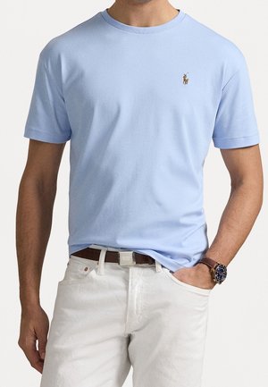 Man draagt lichtblauw T-shirt met korte mouwen en klein geborduurd logo op de borst, witte broek, bruine riem en polshorloge.