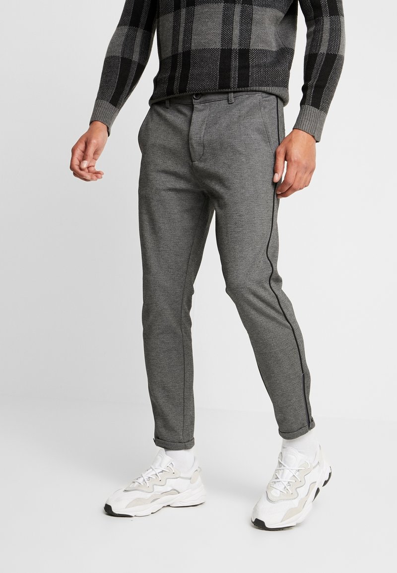 Homme portant un pantalon gris ajusté avec des bandes latérales noires, des baskets blanches et un pull à carreaux gris et noir.