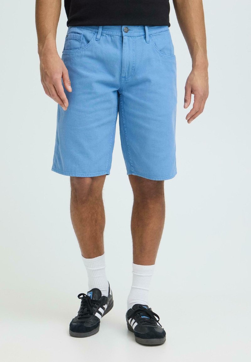 Άνδρας που φοράει ανοιχτό μπλε σορτς μέχρι το γόνατο, λευκές κάλτσες που φτάνουν μέχρι τον αστράγαλο και μαύρα αθλητικά παπούτσια με λευκές ρίγες, στέκεται μπροστά σε ένα απλό φόντο.