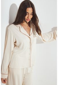 Beige pyjamapak van zachte stof met een knoopsluiting, notch-kraag en contrasterende bruine bies op de mouwen en zak.