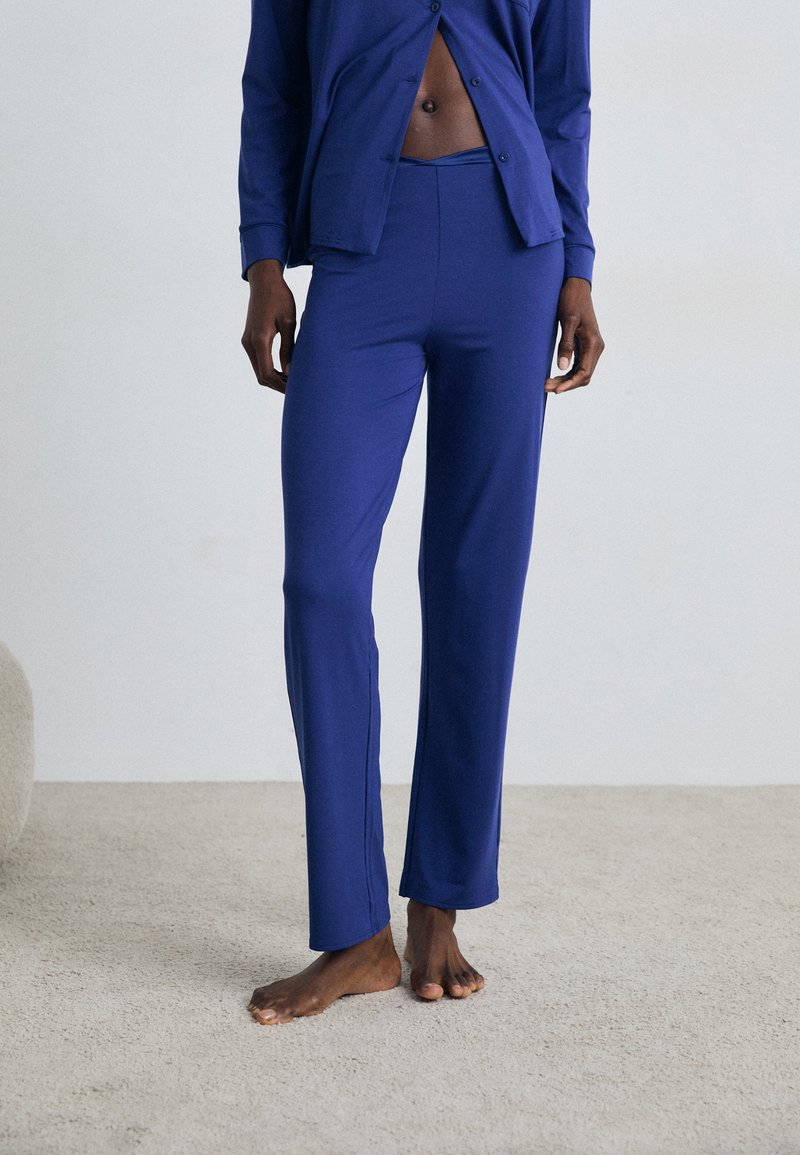 Mėlynas ilgam rankovėmis marškinys su sagomis ir priderintos fitinės kelnės, pagamintos iš minkštos medžiagos. Modelis stovi basi ant šviesaus tekstūruoto paviršiaus.