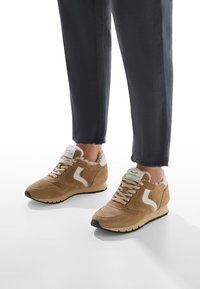 Tan suede sneakers with white accents and faux fur lining, featuring a rounded toe and textured sole. Jeans with frayed hem visible.