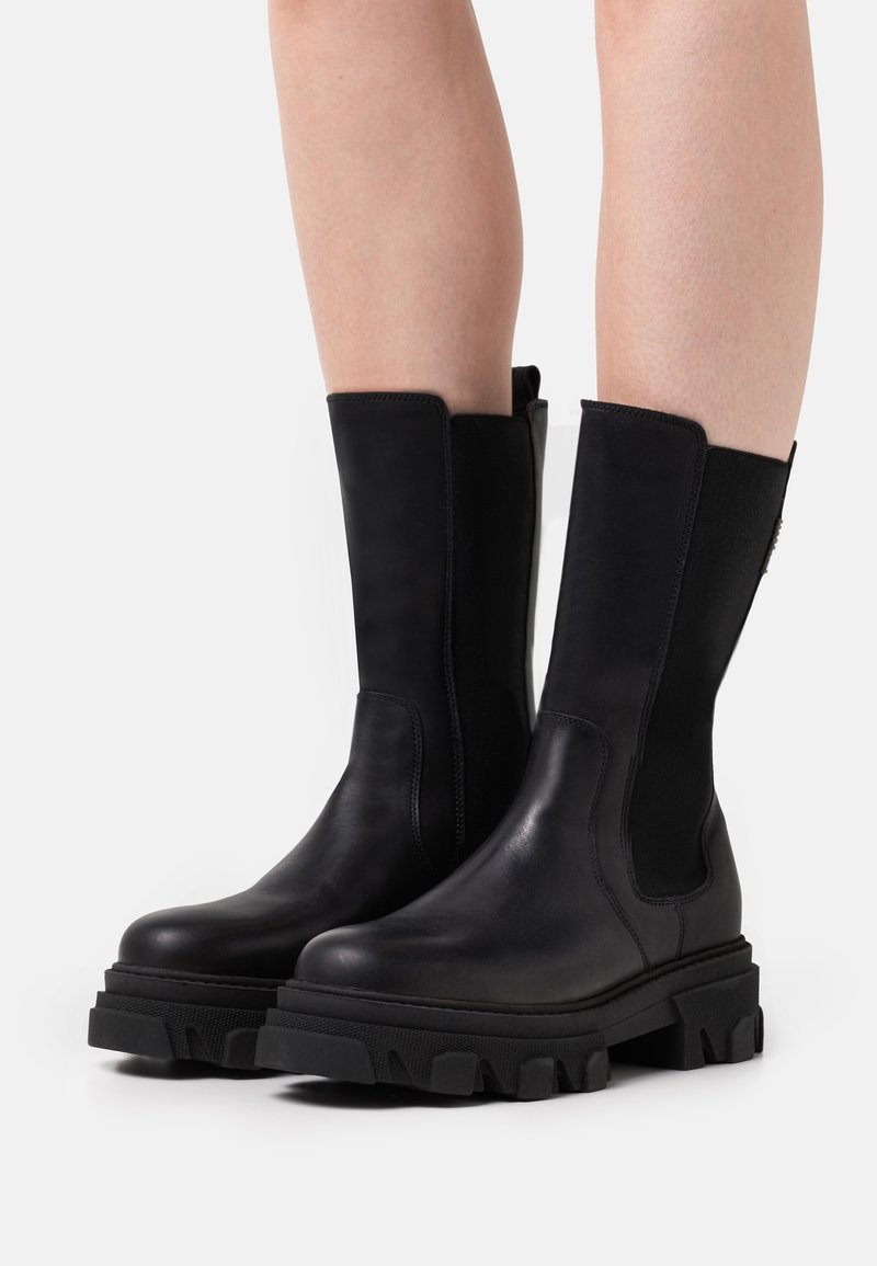 Bottes noires mi-mollet avec semelles épaisses à crampons portées par une personne aux jambes nues sur un fond clair uni.