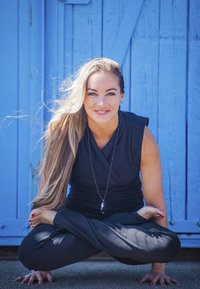 Svart ärmlös topp och byxor, långt hår, sitter i en yogaposition med foten bakom huvudet, ler mot en blå träbakgrund.