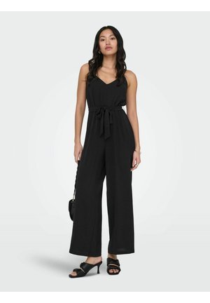 Femme debout portant une combinaison noire sans manches avec une ceinture à la taille, tenant un sac noir et portant des sandales noires à talons.