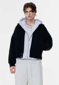 Schwarze, kurz geschnittene Jacke mit einem Frontreißverschluss, über einem hellgrauen Kapuzenpullover getragen. Das Model trägt graue Sweatpants; eine silberne Kette ist sichtbar.