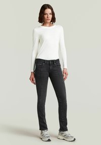 Zwarte jeans met een slanke pasvorm en contrasterende stiksels, gecombineerd met een wit langemouwen shirt. Model draagt grijze sneakers. Eenvoudige achtergrond.